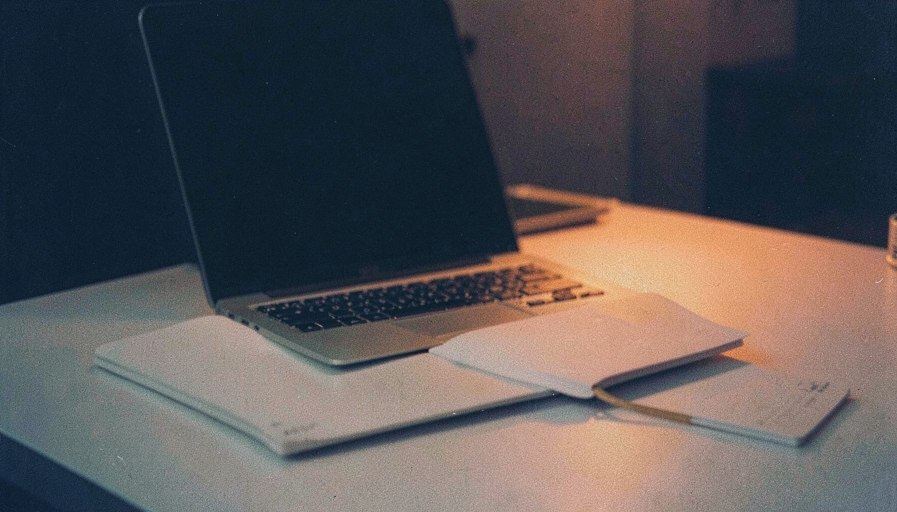 A grainy analog film photograph of an open laptop and notebook on a light grey desk with warm orange accent light, calm editorial mood, deep navy background tones