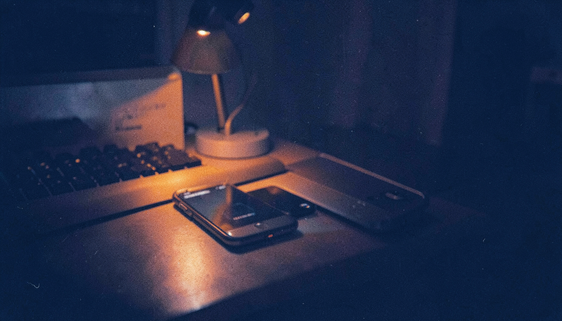 A grainy analog film photograph of a desk scene with a smartphone and laptop glowing softly in deep navy blue shadow, warm orange lamp light spilling across the frame, calm and focused mood