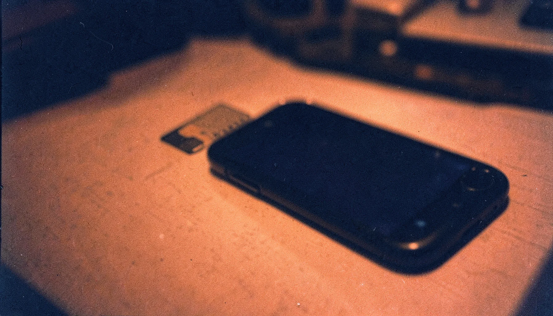 A grainy analog film photograph of a sim card and smartphone on a desk under warm orange light with deep navy shadows, calm technical mood