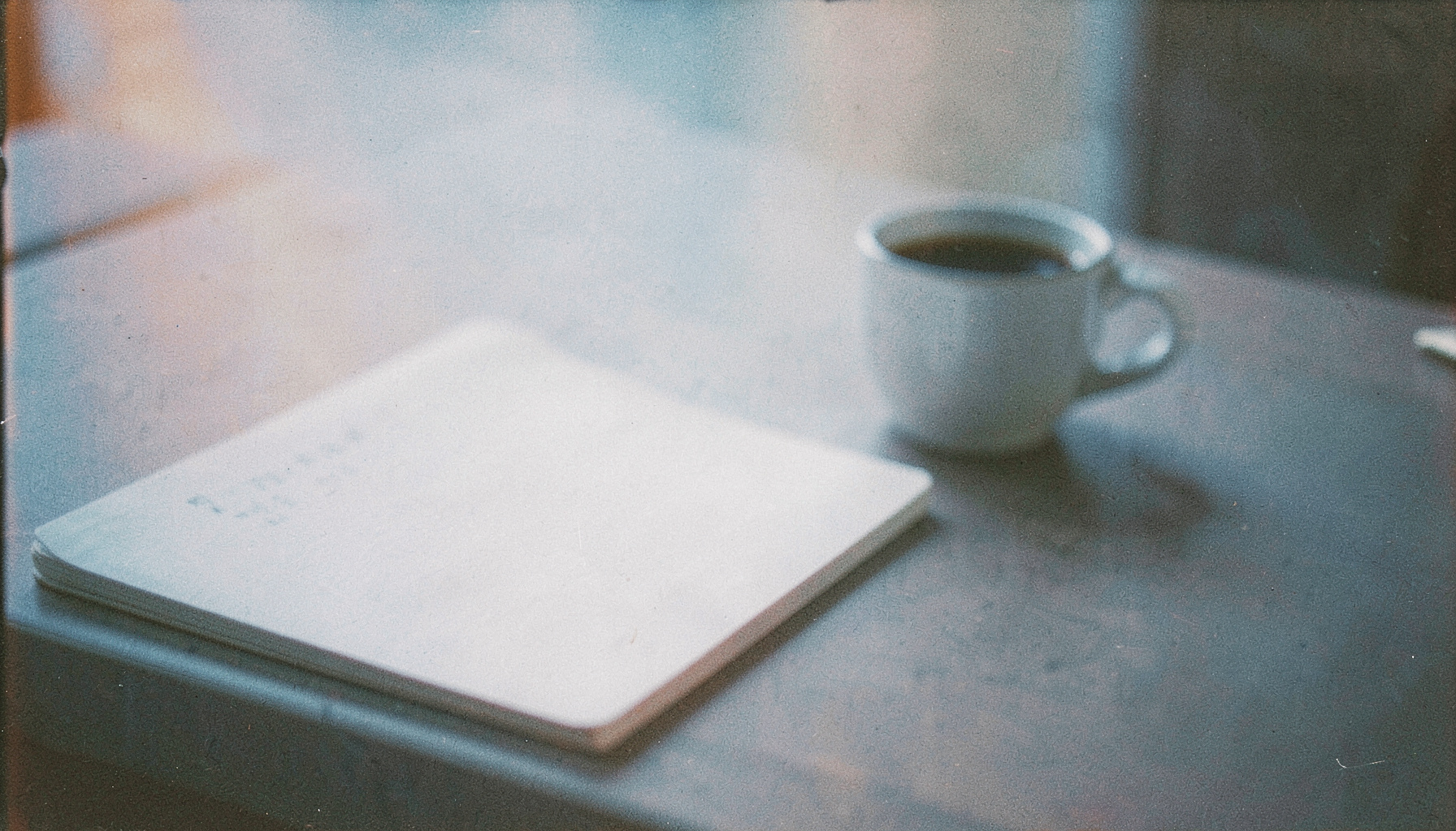 A muted analog film photograph of a flat desk surface with a notebook and a coffee cup, soft blue tones and visible grain, calm and editorial mood.