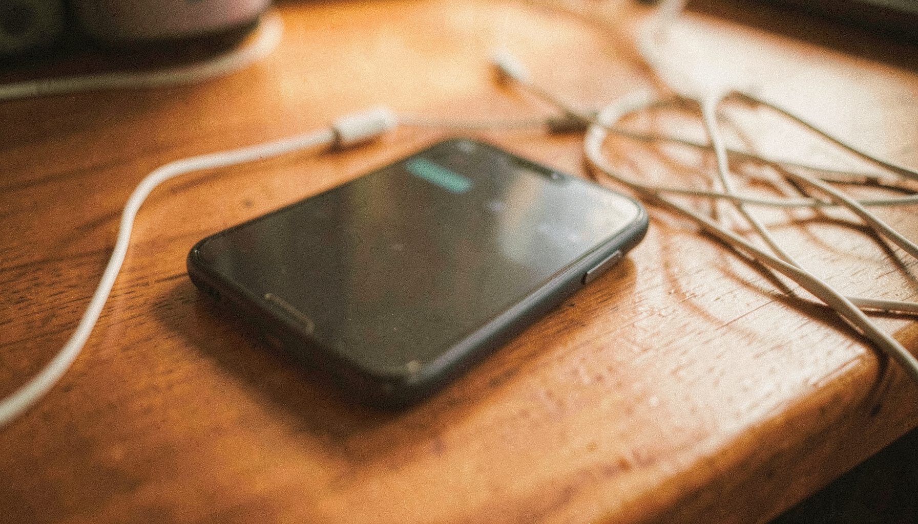 Close-up analog photograph of a smartphone resting on a wooden desk beside tangled earbuds, soft morning light with visible grain and warm amber tones