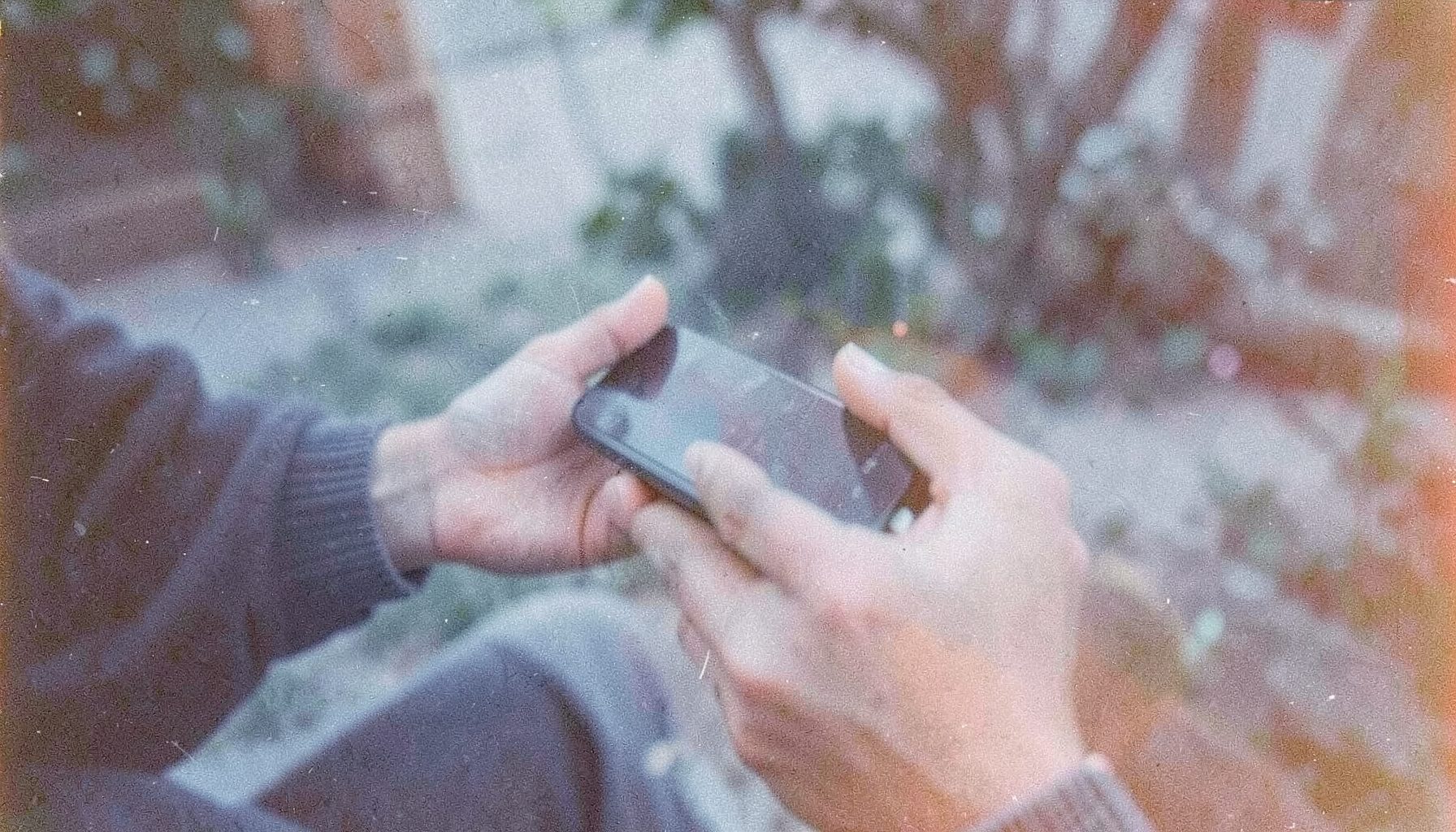 Faded analog photograph of a pair of hands holding a smartphone outdoors, cool blue and gray tones with soft grain