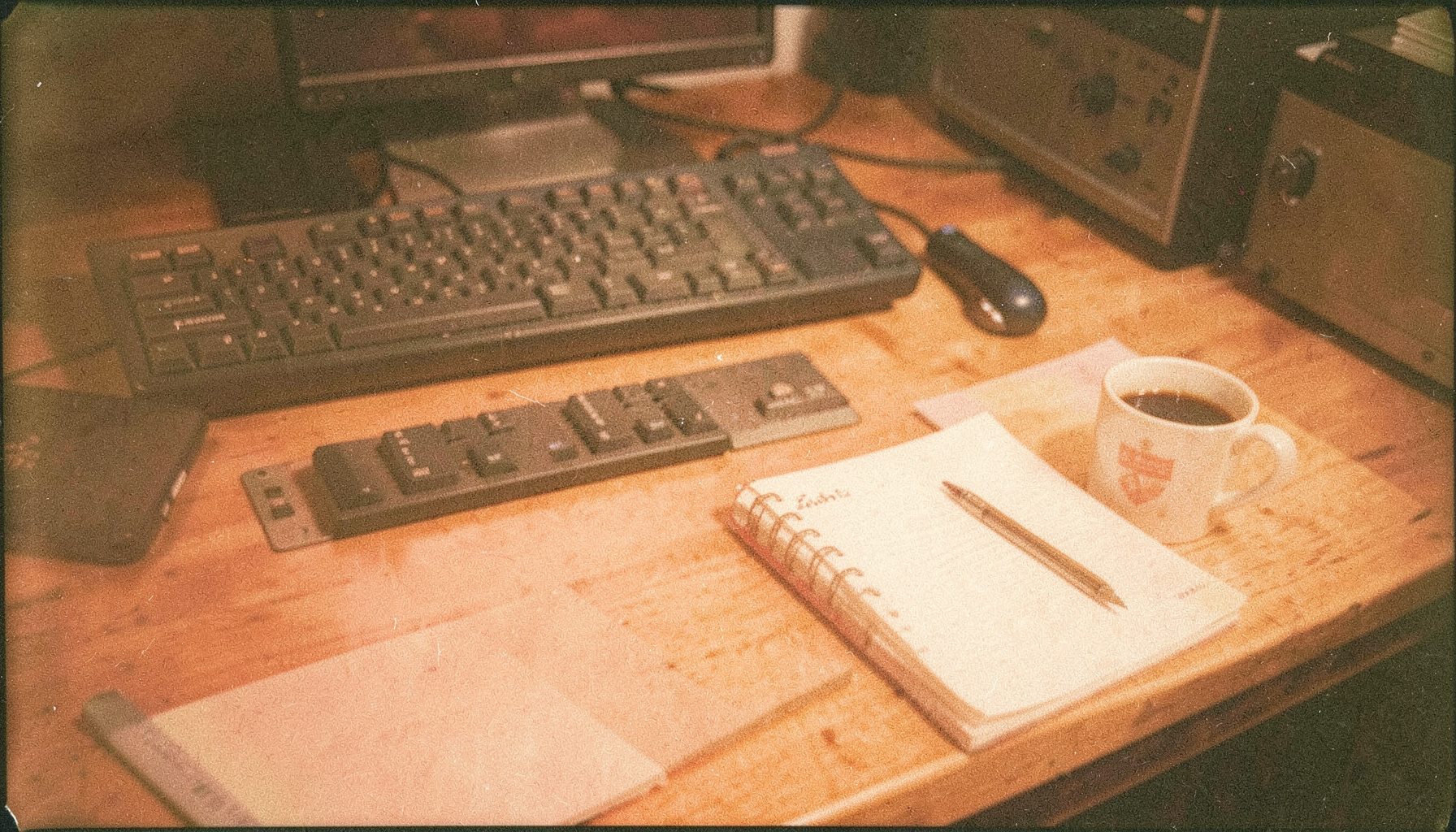 Analog film photograph of a developer desk with a keyboard, notebook, and coffee mug on warm-toned wood, soft grain and amber light, calm and focused mood